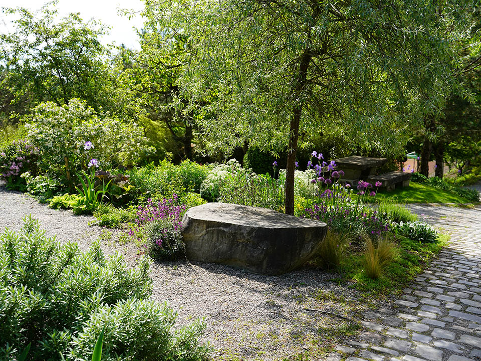 Gartengestaltung mit Bäumen und Sträucher und einen Naturstein als Sitzplatzmöglichkeit