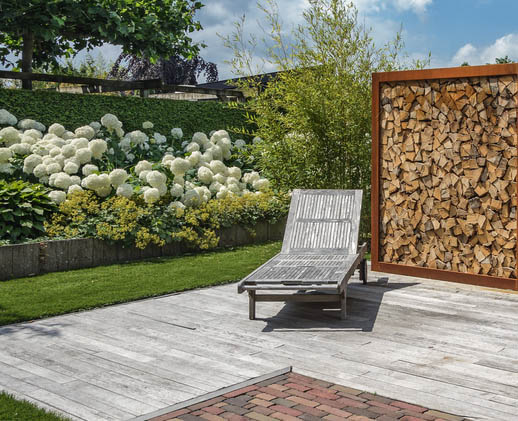 Terrasse mit Liegestuhl und gestapeltem Holz und Stauden im Hintergrund